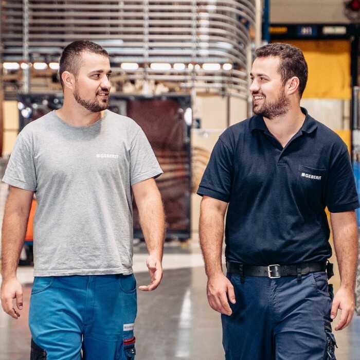 Two workers walking and talking in a factory, surrounded by equipment and storage containers.