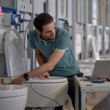 A man in a teal shirt is examining a toilet with a laptop and other equipment.