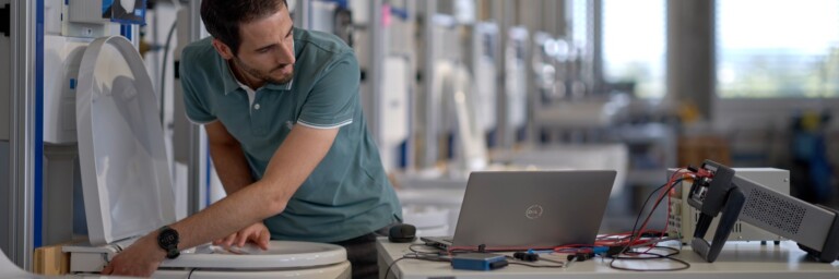 A man inspects a toilet with a laptop and testing equipment on the table.