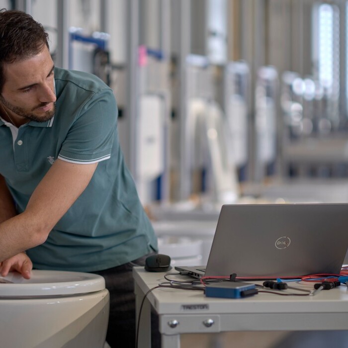 A man inspects a toilet with a laptop and testing equipment on the table.