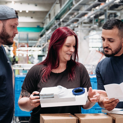 Drei Personen in Arbeitskleidung prüfen gemeinsam ein Gerät und eine Anleitung in einer Produktionsumgebung.