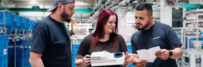 Drei Personen in Arbeitskleidung prüfen gemeinsam ein Gerät und eine Anleitung in einer Produktionsumgebung.