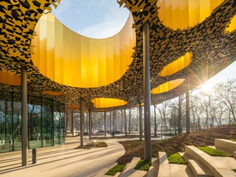 Moderner Aussenbereich mit auffälligen gelben, gewölbten Überdachungen und Glasfassade, umgeben von Bäumen.