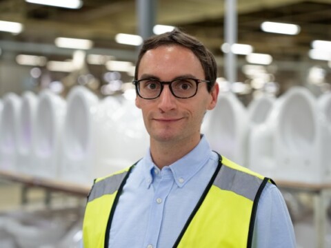 A man wearing glasses and a safety vest stands in a factory with rows of white toilets in the background.