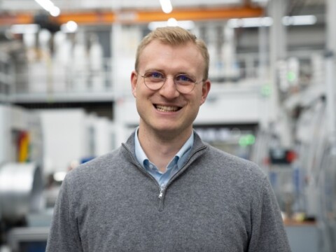 A smiling man in a gray sweater stands in a factory setting, with machines and industrial equipment in the background.