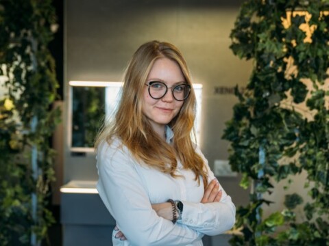 A young woman with long blonde hair and glasses stands confidently with crossed arms in a modern, greenery-filled interior.