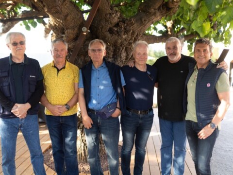 Sechs Männer stehen lächelnd in einer Reihe unter einem grossen Baum.