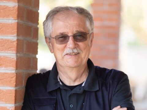 Ein älterer Mann mit Brille und grauen Haaren steht lächelnd an einer ziegelsteinernen Säule.