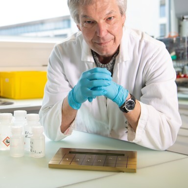 A scientist in a lab coat and blue gloves is leaning over a table with containers and a metal in a bright laboratory setting.