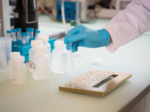 A gloved hand holding a pipette over a metal, with several clear and blue laboratory bottles in the background.