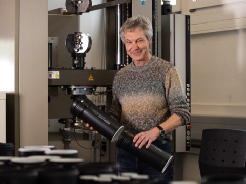 A smiling man holding a large cylindrical object in a laboratory setting with research equipment in the background.