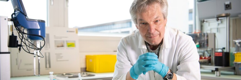 A man in a lab coat and blue gloves is working at a laboratory bench surrounded by various lab equipment and containers.