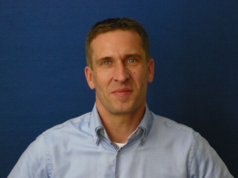A man with short hair wearing a light blue shirt, smiling against a dark blue background.