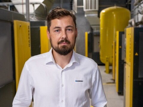 A man in a white shirt stands in an industrial setting with yellow machinery in the background.