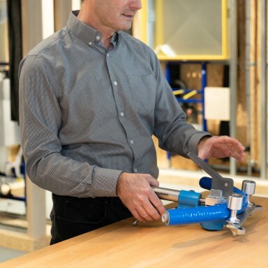 Ein Mann in einem grauen Hemd benutzt ein Werkzeug auf einem Holztisch in einer Werkstatt.