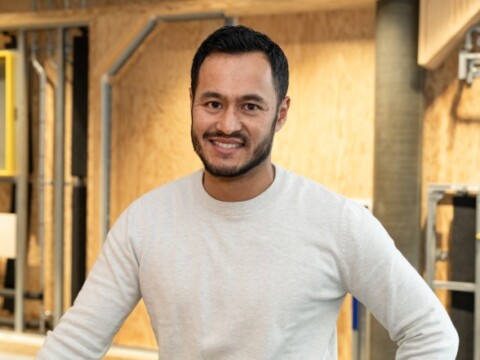 A man smiling dressed in a light grey sweater and standing in an industrial-style setting with wooden walls and visible pipes.
