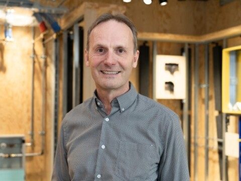 A smiling man in a button-up gray shirt stands in a modern space with wooden walls and visible pipes.