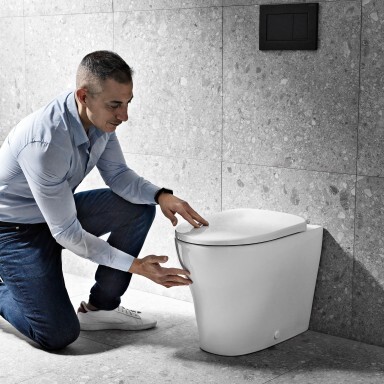 Mann kniet neben einer modernen, weissen Toilette und überprüft sie in einem Badezimmer mit grauen Wänden.