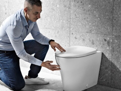 Ein Mann in einem Hemd kniet neben einer modernen weissen Toilette und untersucht sie.