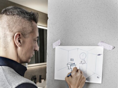 Ein Mann zeichnet an einer Wand eine skizzenhafte Darstellung eines Badezimmers.