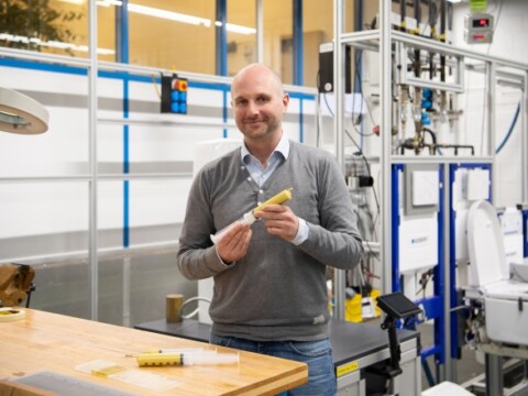 Ein Mann hält eine gelbe Spritze in einer industriellen Umgebung mit Maschinen im Hintergrund.