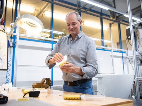 Ein Mann mit grauem Hemd, der an einem Tisch in einem modernen Arbeitsraum eine gelbe Komponente bearbeitet.