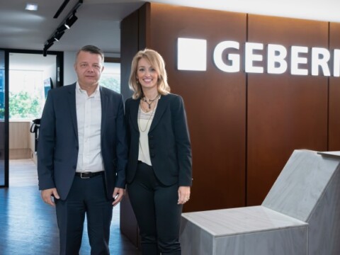 Geschäftsleute stehen lächelnd im Büro vor einem beleuchteten Logo mit dem Namen "GEBERIT".