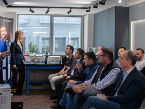 Eine Gruppe von Menschen sitzt in einem modernen Raum und hört einer Präsentation zu, während eine Frau vorne steht und spricht.