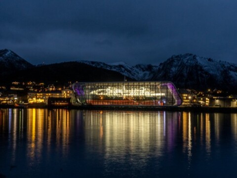 Ein beleuchtetes modernes Gebäude am Wasser bei Nacht, umgeben von schneebedeckten Bergen und einer Stadt mit warmem Licht reflektiert im Fluss.