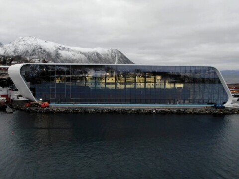 Moderne, futuristische Gebäude am Wasser mit einer grossen Glasfassade und schneebedeckten Bergen im Hintergrund.