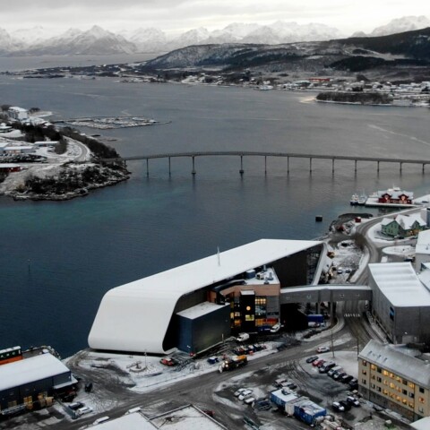 Luftaufnahme einer schneebedeckten Küstenstadt mit einem Fjord, einer Brücke und Bergen im Hintergrund.