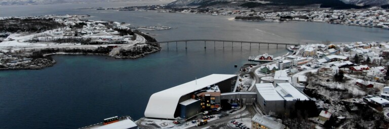 Luftaufnahme einer schneebedeckten Küstenstadt mit einem Fjord, einer Brücke und Bergen im Hintergrund.