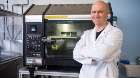 Ein Mann in einem Laborkittel steht mit verschränkten Armen neben einem grossen wissenschaftlichen Gerät in einem Labor.