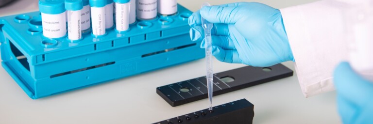 Ein Laborgestell mit Reagenzgläsern und einer Hand mit blauem Handschuh, die eine Pipette mit Flüssigkeit über einen Teststreifen hält.