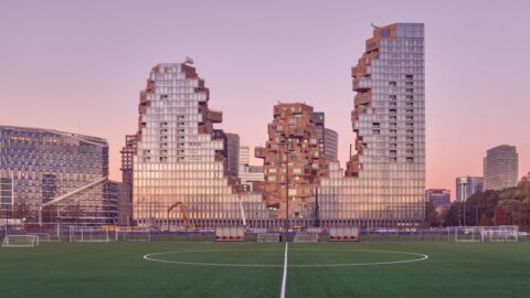 Modern skyscrapers with fragmented architecture, set against a twilight sky, viewed from a sports field.