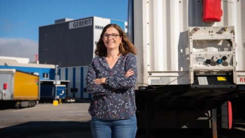 A woman with long brown hair stands confidently with arms crossed in front of a lorry and warehouse, smiling in an industrial setting.