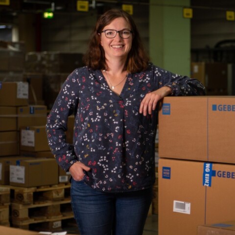 A woman stands smiling next to stacked cardboard boxes in a warehouse setting.