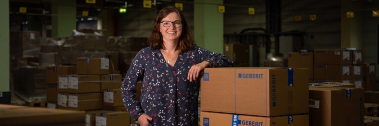 A woman stands smiling next to stacked cardboard boxes in a warehouse setting.