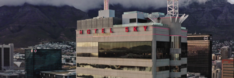 A modern high-rise building labeled "Hotel Sky," surrounded by urban structures and mountains in the background.