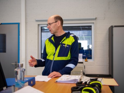 Ein Mann im blauen und grünen Arbeitsanzug steht an einem Tisch, während er mit einer Hand gestikuliert und Papiere vor ihm liegen.