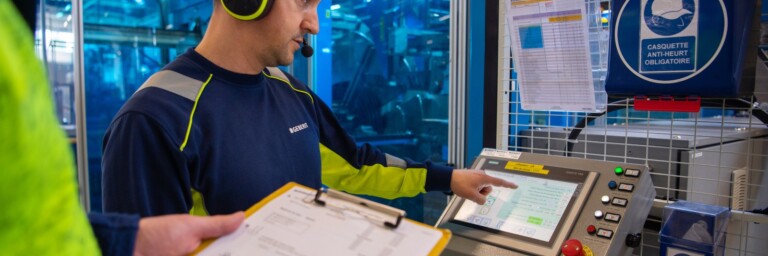 Ein Mann in Arbeitskleidung mit Kopfhörern steht vor einem Kontrollpaneel und berührt den Bildschirm.