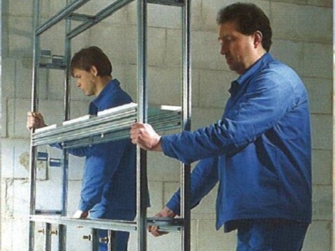 Ein Männer in blauer Arbeitskleidung arbeiten an einem Metallrahmen in einem Raum mit Wänden aus Betonsteinen.