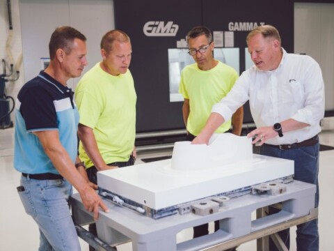 Vier Männer betrachten gemeinsam ein weisses Objekt auf einem Tisch in einer modernen Werkstatt.