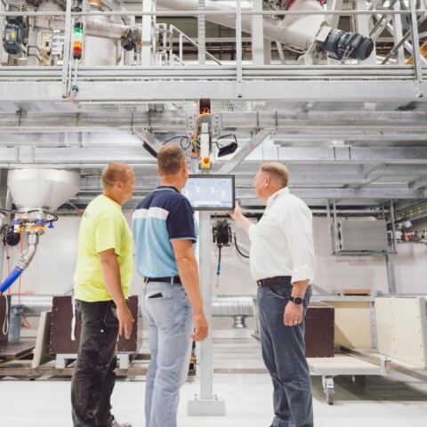 Three men looking at a screen in a large industrial space, with metal frameworks and machinery in the background.