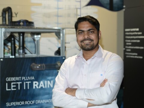 Ein lächelnder Mann mit kurzem, dunklem Haar steht mit verschränkten Armen vor einem Ausstellungsstand, der Produkte zur Dachentwässerung zeigt.