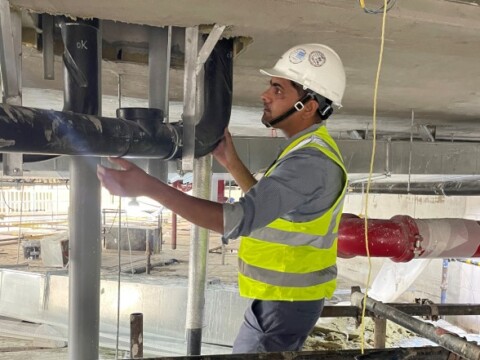 Ein Mann in einem Sicherheitshelm und einer Warnweste, der an Rohren auf einer Baustelle arbeitet.