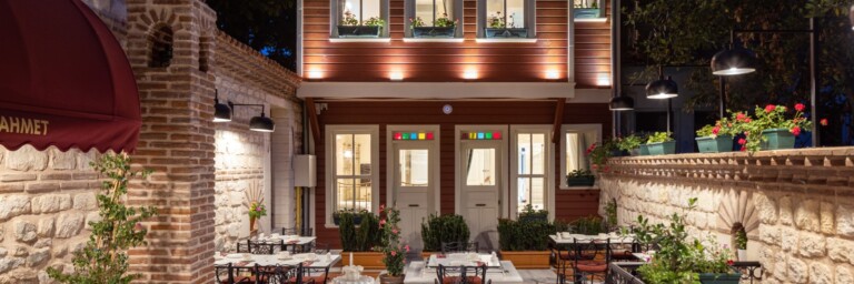 A beautifully illuminated hotel exterior featuring a wooden building with large windows, surrounded by tables and greenery in a cozy night setting.
