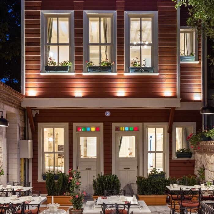 A beautifully illuminated hotel exterior featuring a wooden building with large windows, surrounded by tables and greenery in a cozy night setting.