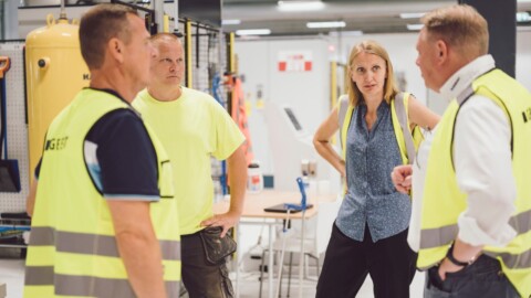 Vier Personen in Warnwesten stehen in einem modernen Arbeitsumfeld und diskutieren miteinander.