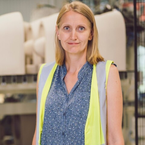 Eine Frau mit einem blauen Oberteil und einer gelben Warnweste steht vor industriellen Maschinen in einer Fabrik.